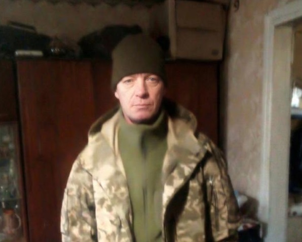 Чоловік у камуфляжній куртці, зеленому светрі та шапці стоїть у приміщенні, дивиться прямо в камеру.