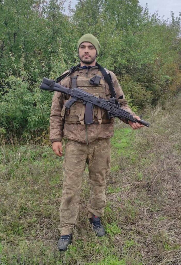 Чоловік у військовій формі та з автоматом в руках стоїть у полі.