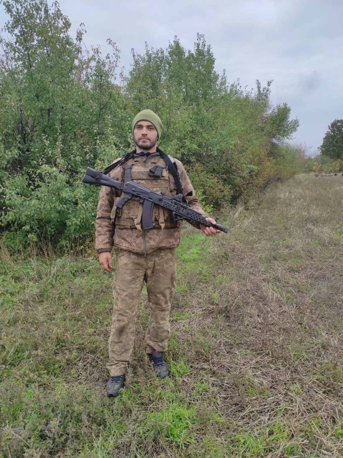 Чоловік у військовій формі та з кулеметом в руках стоїть у полі.