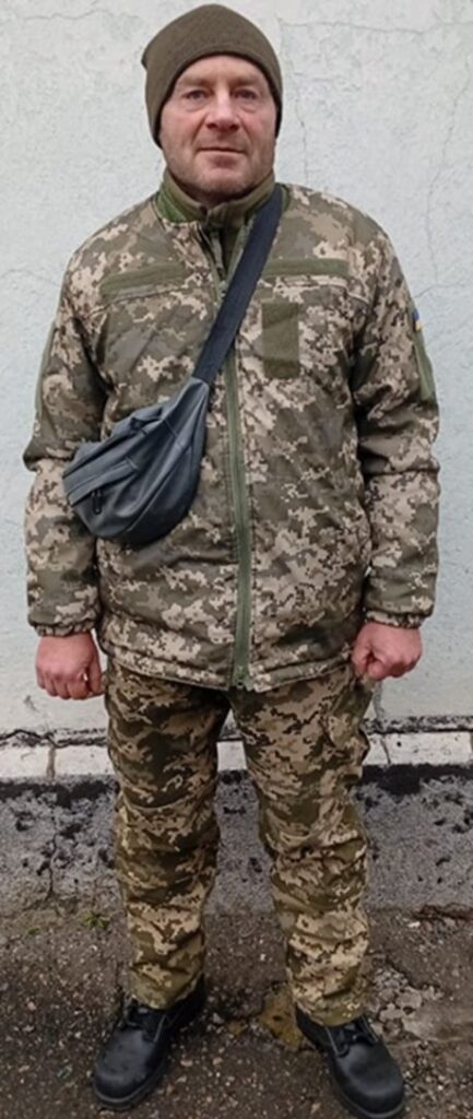 Чоловік у камуфляжній формі та з чорною сумкою через плече стоїть біля сірої стіни.