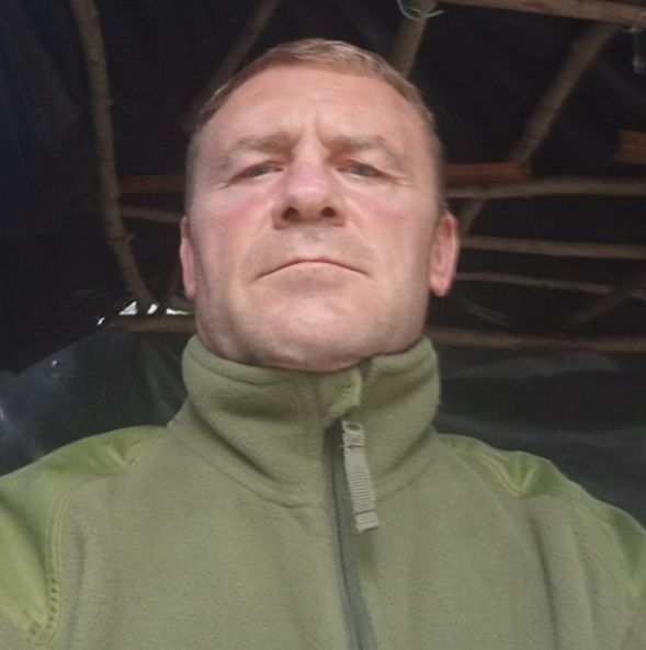 Чоловік у світло-зеленому флісовому піджаку з високим коміром дивиться прямо в камеру.