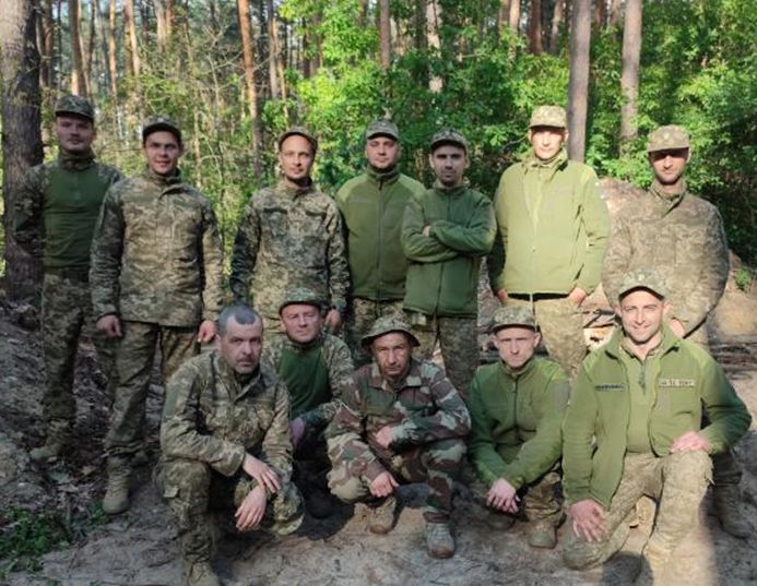 Група військових у камуфляжній формі, частина з яких озброєна, позирують на тлі лісу.