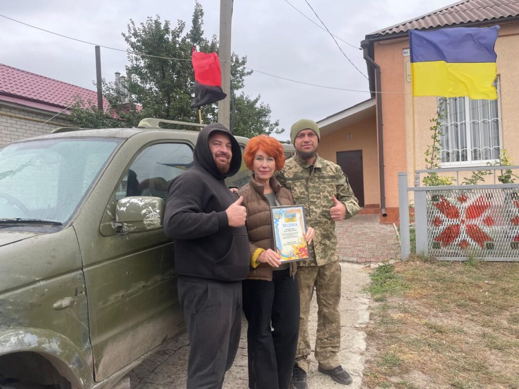 Чоловік у чорному худи, жінка із сертифікатом та чоловік у камуфляжі позують біля зеленого пікапа.