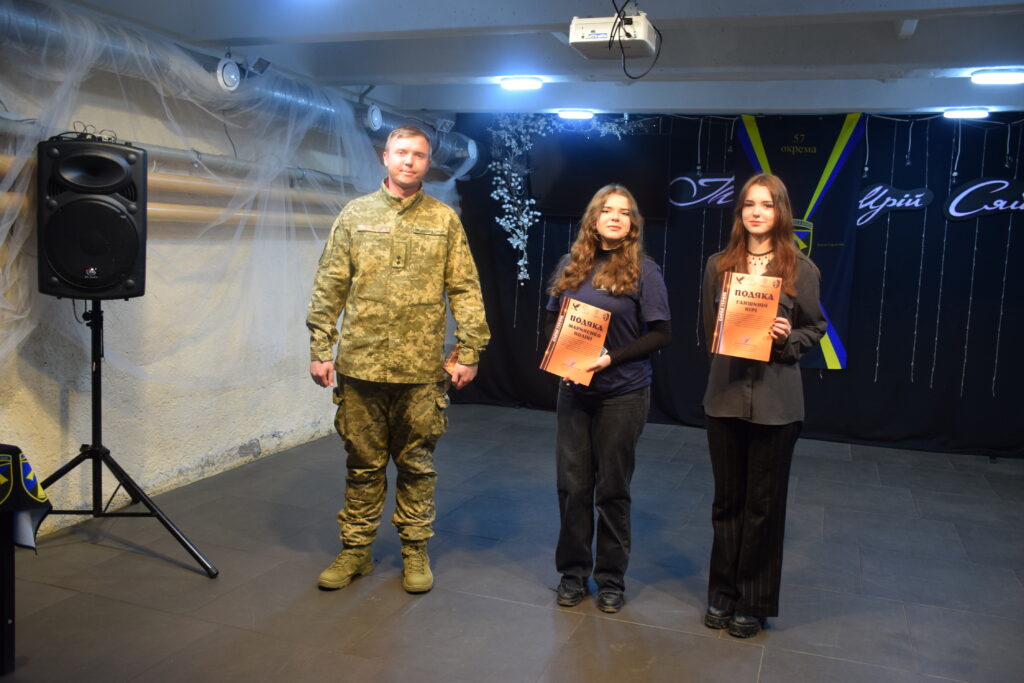 Чоловік у військовій формі стоїть поряд із двома дівчинам, які тримають у руках помаранчеві документи.