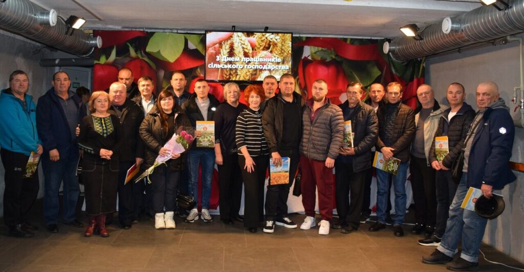 Кілька людей стоять у приміщенні, більшість тримають у руках журнали, жінка посередині тримає букет квітів, позаду них екран із текстом українською мовою та зображенням колосків.
