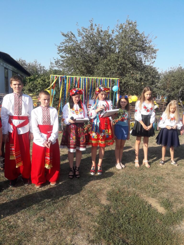 Група дітей у національних українських костюмах стоїть на галявині біля будинку з різнобарвними стрічками позаду.
