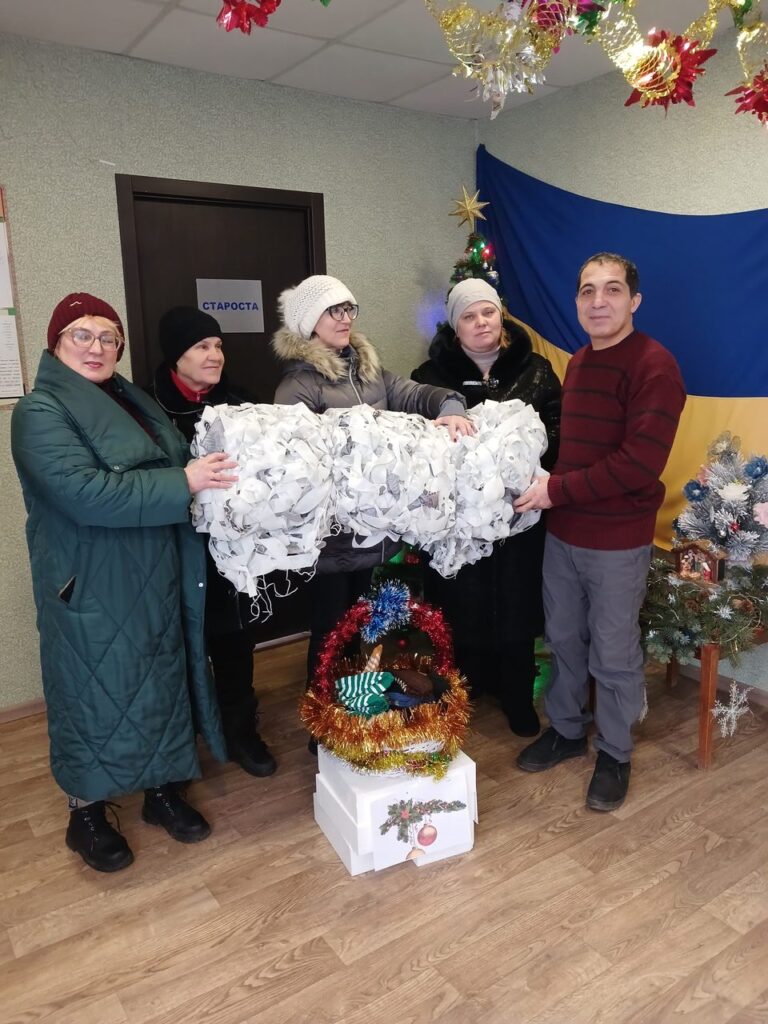 П'ять людей тримають велику кількість зібраних білих паперових зірок, навколо різдвяні прикраси.