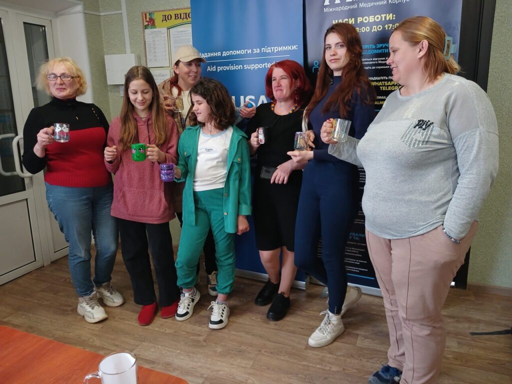 Жінки та дівчата стоять біля банера організації, тримаючи в руках розфарбовані чашки.