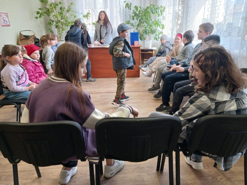 Діти сидять у колі на стільцях, один хлопчик стоїть усередині кола і щось розповідає іншим.