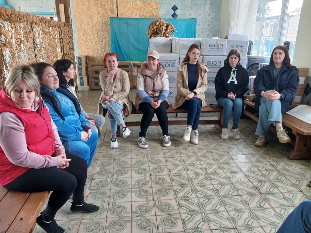 Вісім жінок сидять на лавках у приміщенні з коробками та різними предметами позаду них.