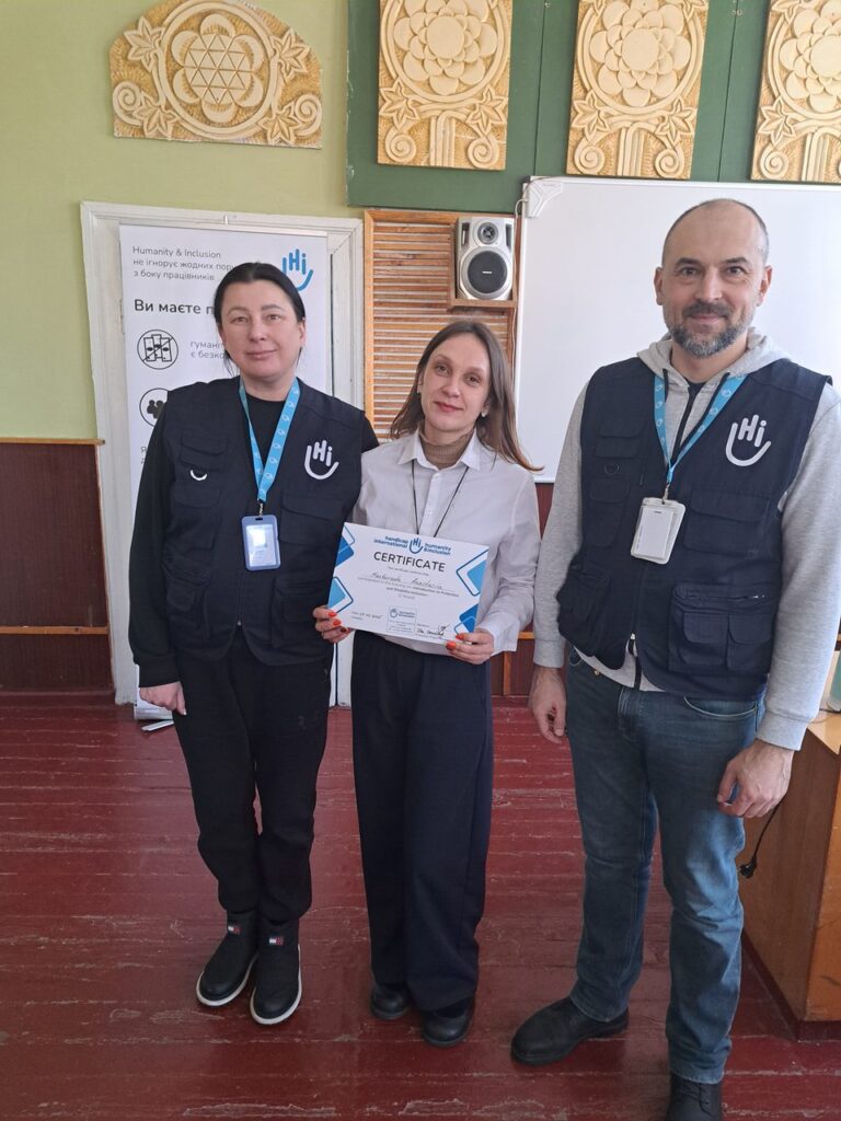 Жінка тримає сертифікат та стоїть між двома чоловіками у чорних жилках із логотипом, усі мають ідентифікаційні бейджи на шиї.