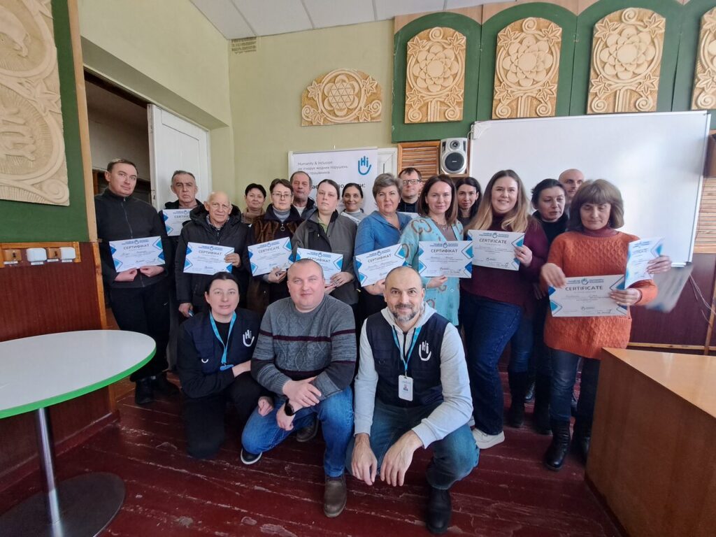 Кілька людей різних статей та віку тримають сертифікати й позують у приміщенні із зеленою стіною та дерев’яними декоративними елементами.
