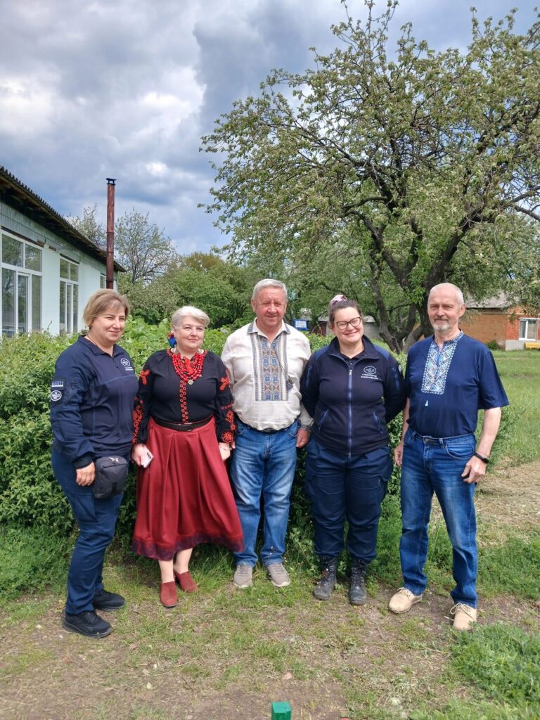 П'ять осіб стоять поряд на галявині біля будинку, серед них дві жінки у темних куртках та троє у вишиванках і святкових нарядах.