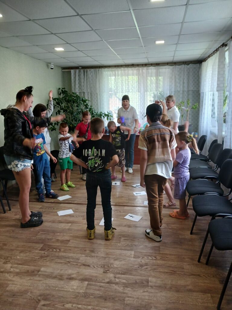 Діти та дорослі стоять у колі, виконуючи завдання на підлозі із паперів у приміщенні з дерев'яною підлогою та білими шторами на вікнах.