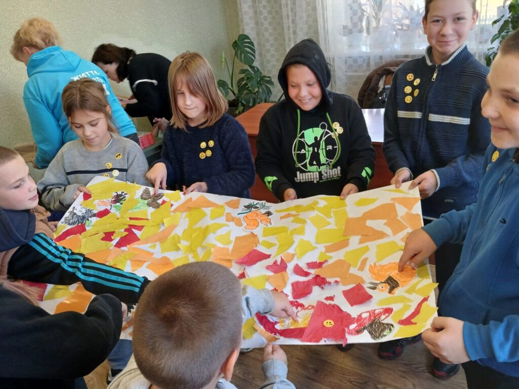 Діти тримають велике кольорове паперове зображення осіннього пейзажу з елементами малюнків.