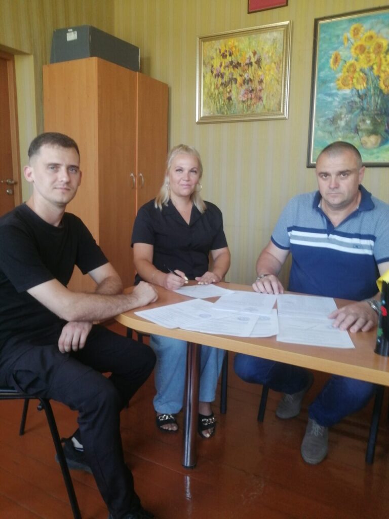 Чоловік у чорному сидить біля столу з документами разом із жінкою у чорному та чоловіком у блакитному, розглядаючи папери.