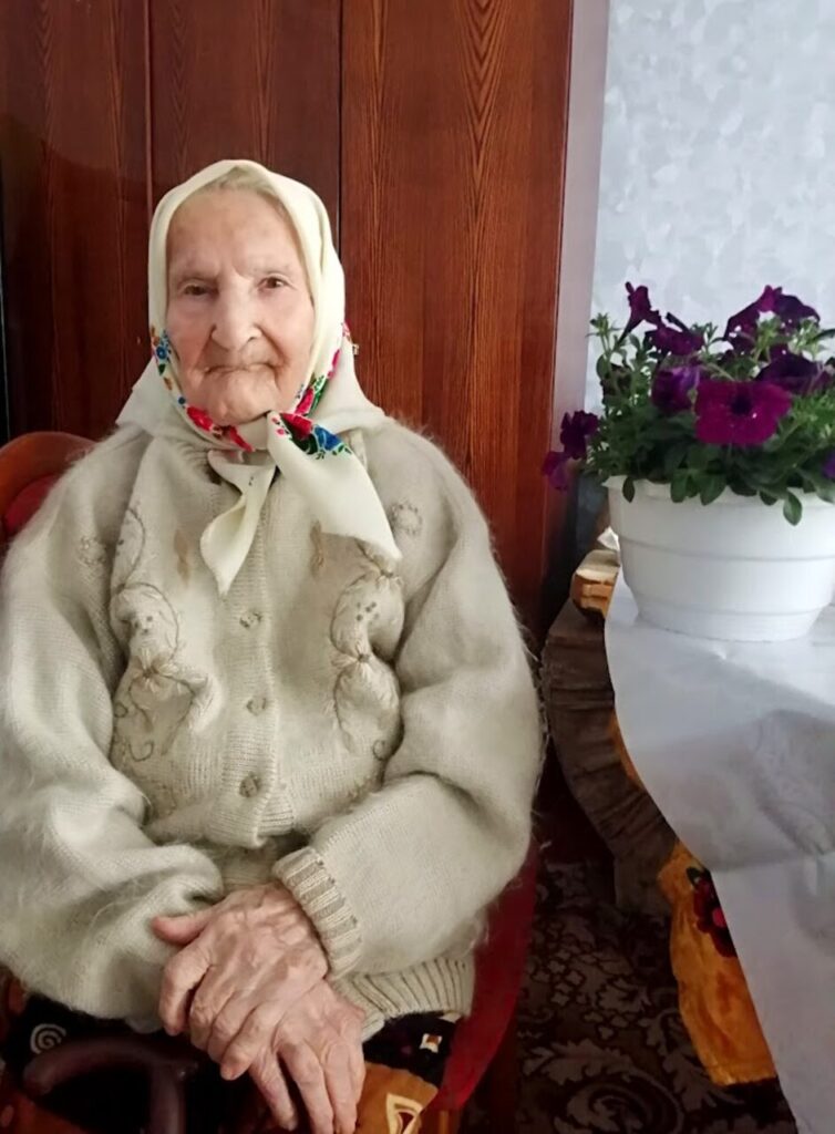 Літня жінка у світлому одязі та хустці сидить у кріслі біля столу з квітами.