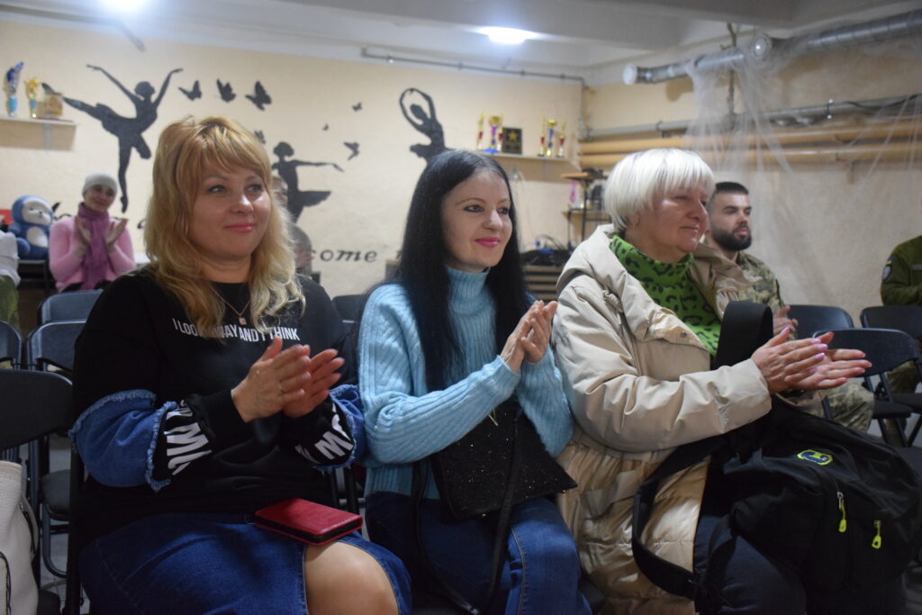 Три жінки сидять у ряді стільців і аплодують, поряд із ними сидить чоловік у військовій формі.