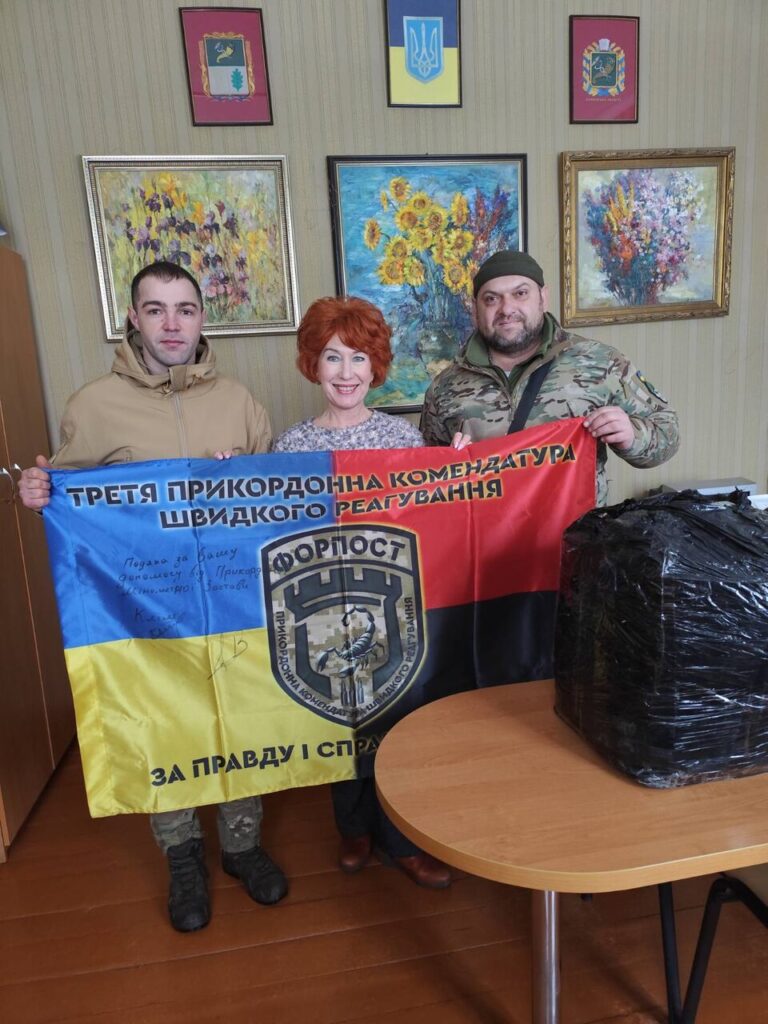 Чоловік у камуфляжі та жінка тримають прапор із емблемою та написом українською мовою, поряд інший чоловік у куртці.