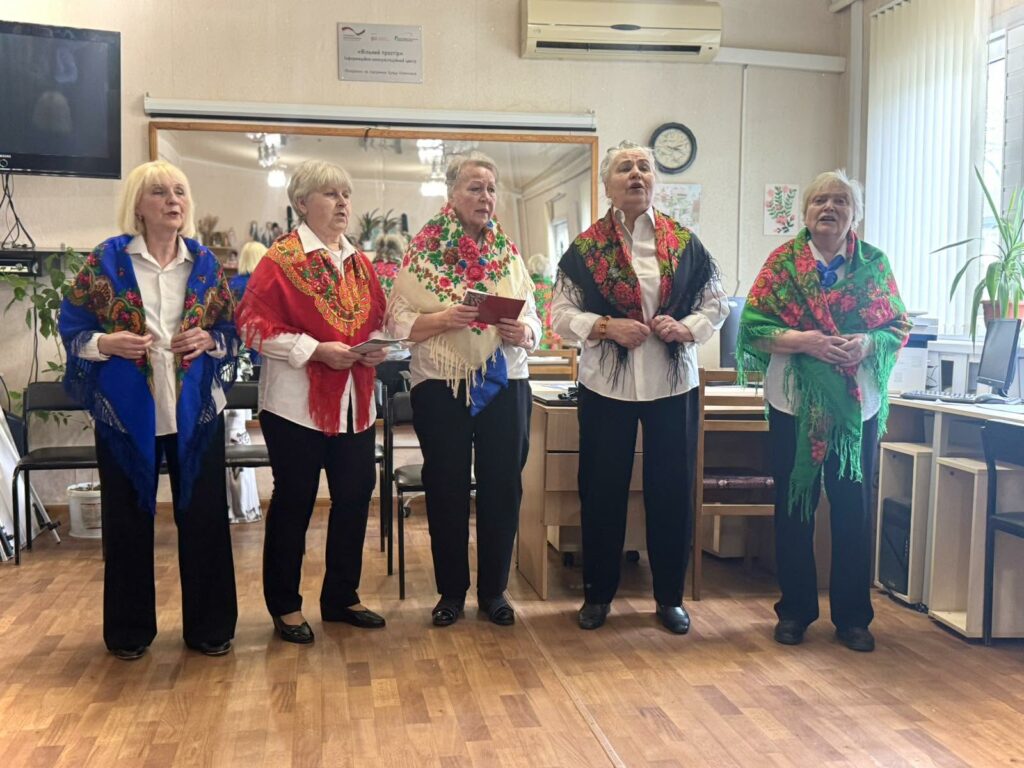 П'ять жінок співають у приміщенні, одягнені у вишиті кольорові шалі та білі сорочки.