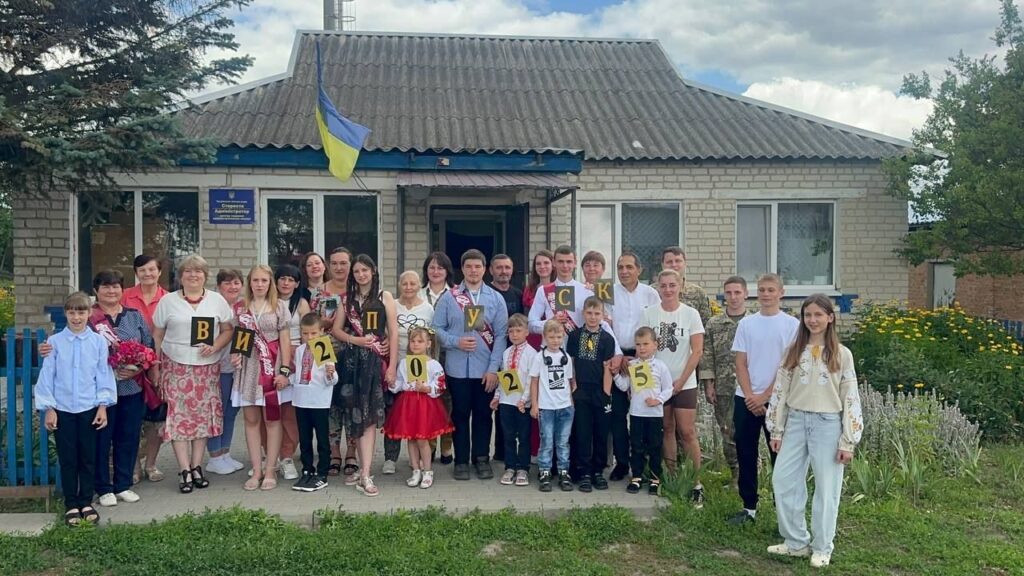 Велика група людей різного віку стоїть перед будівлею з українським прапором, деякі тримають літери, що складають напис