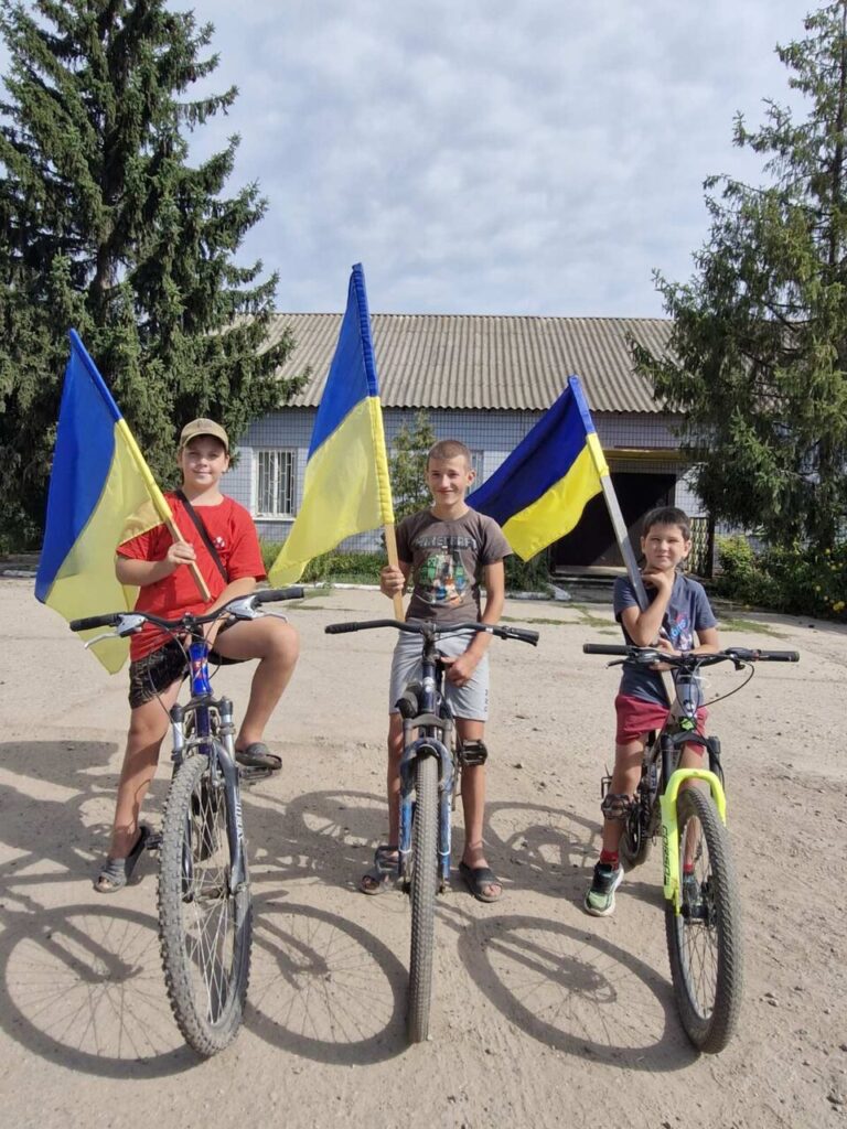 Троє хлопців підліткового віку позують із українськими прапорами, сидячи та стоячи поруч із велосипедами на піщаній дорозі біля будинку.