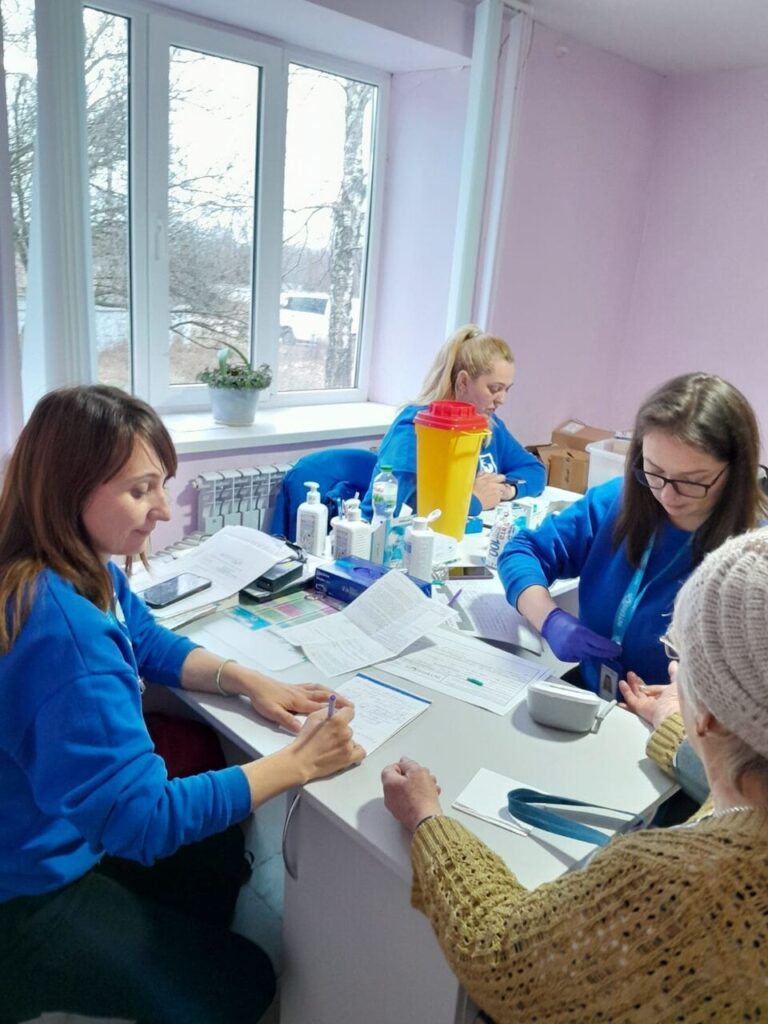 Жінка у синьому одязі та окулярах пише на папері, сидячи за столом разом із жінкою у жовтому одязі; інша жінка у синьому дивиться на їхні дії; третя жінка у синьому працює на тлі.