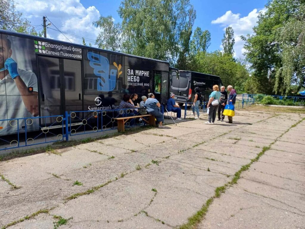 Лікаря в синіх гумових рукавичках тримає біля обличчя пацієнта прилад для забору біологічного матеріалу, на великому автобусі із написом українською мовою