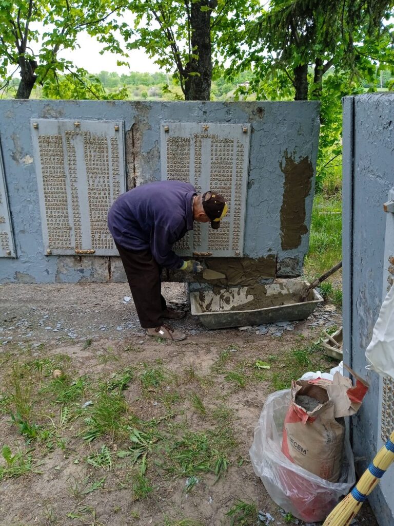 Чоловік у фіолетовій куртці та чорній шапці з жовтим значком ремонтує стару пам'ятну плиту із написами російською мовою.
