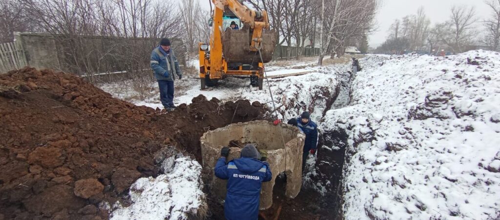 Чоловік у формі встановлює бетонну кільцеву секцію у викопану траншею під снігом, поруч робітники та техніка.