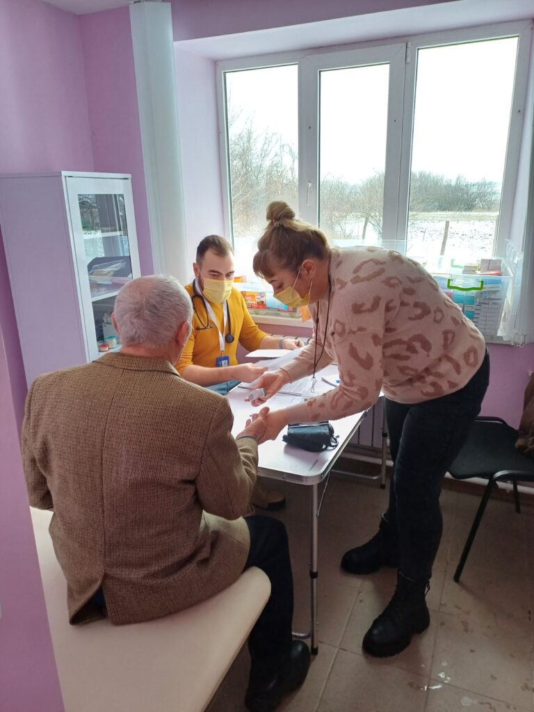Лікар у жовтому одязі та жінка в рожевому одязі проводять медичну процедуру чоловікові із сідим волоссям, сидячому на лавці.
