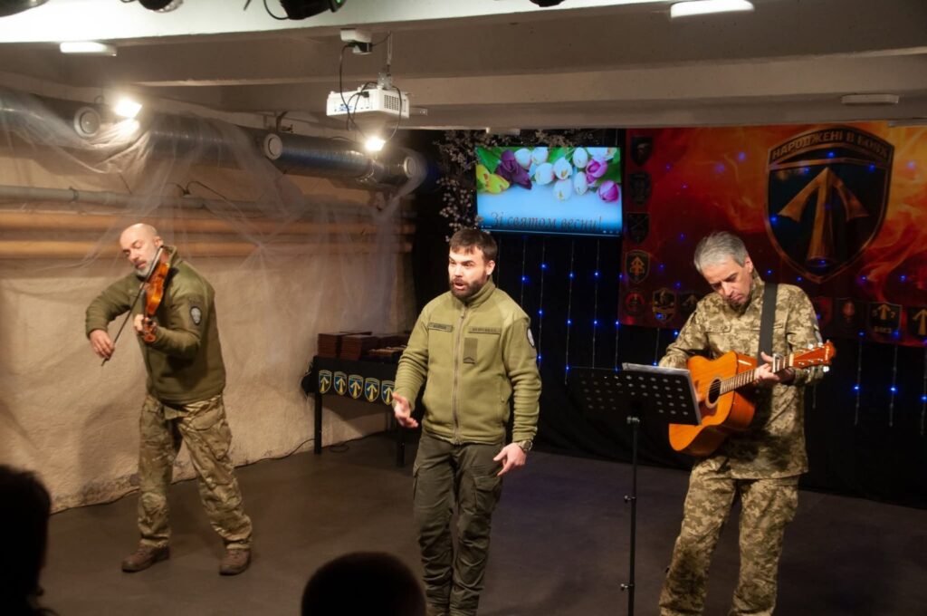Три військових виступають на сцені: один грає на скрипці, інший співає, а третій грає на гітарі.