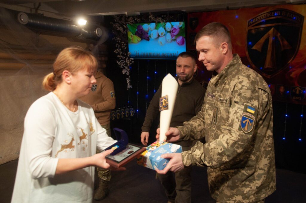 Чоловік у військовій формі вручає жінці подарунки, двоє інших людей стоять позаду них.