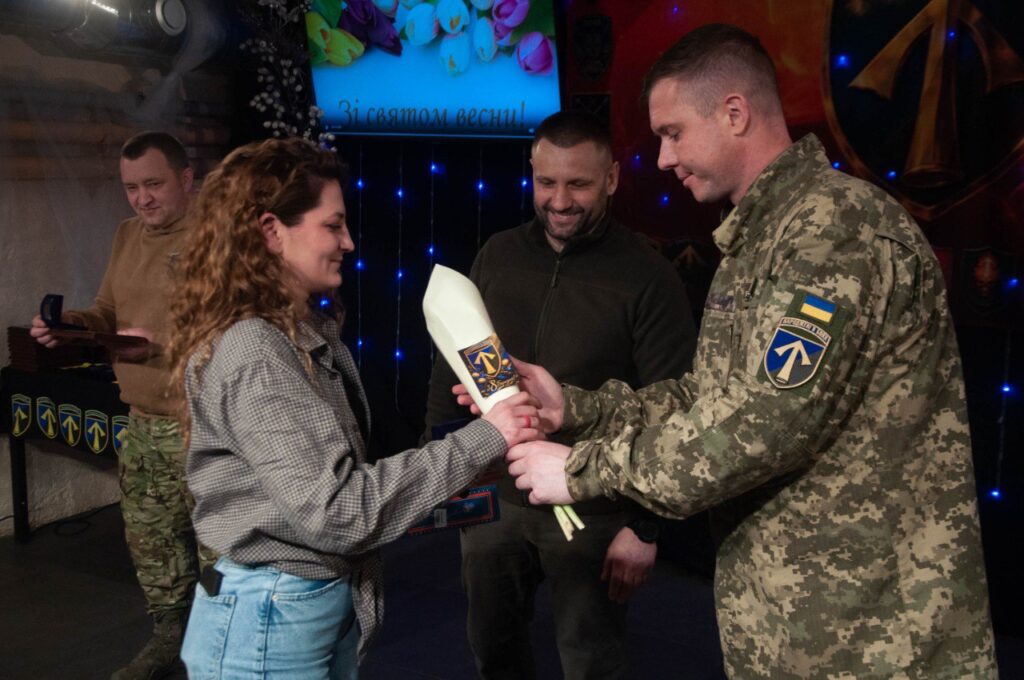 Чоловік у військовій формі вручає жінці велику білу пляшку з етикеткою, на якій розміщений логотип із стрілою, у приміщенні з іншими людьми на тлі.