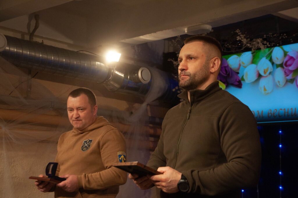 Два чоловіки тримають дерев’яні підноси, один у коричневому худи, інший у темно-зеленому флісовому дресі.