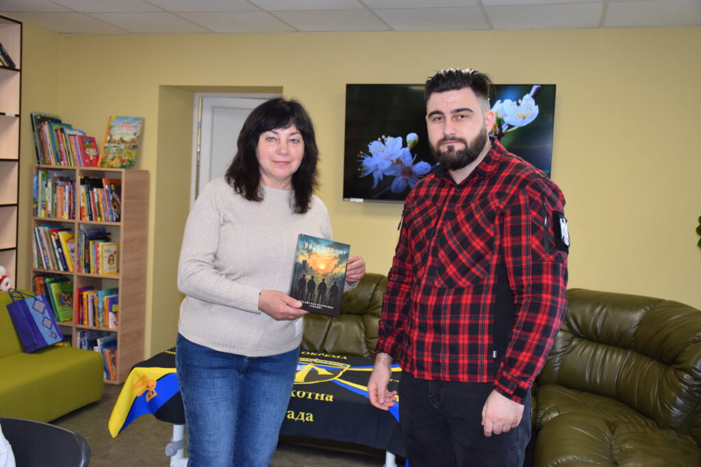 Жінка та чоловік стоять у кімнаті, жінка тримає книгу із зображенням людей на обкладинці.