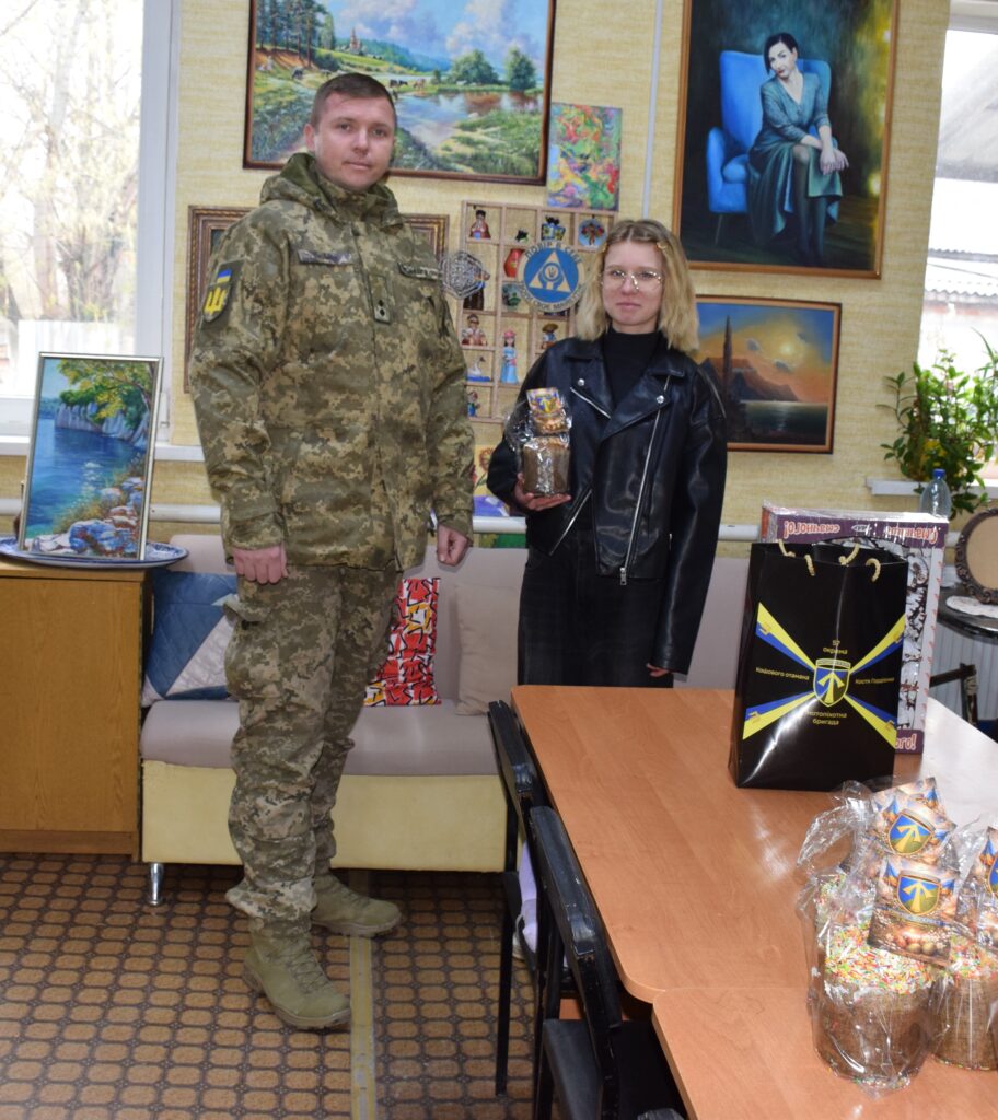 Чоловік у військовій формі та жінка у чорній шкіряній куртці стоять поруч із таблицею, на якій лежать пакети зі солодощами.