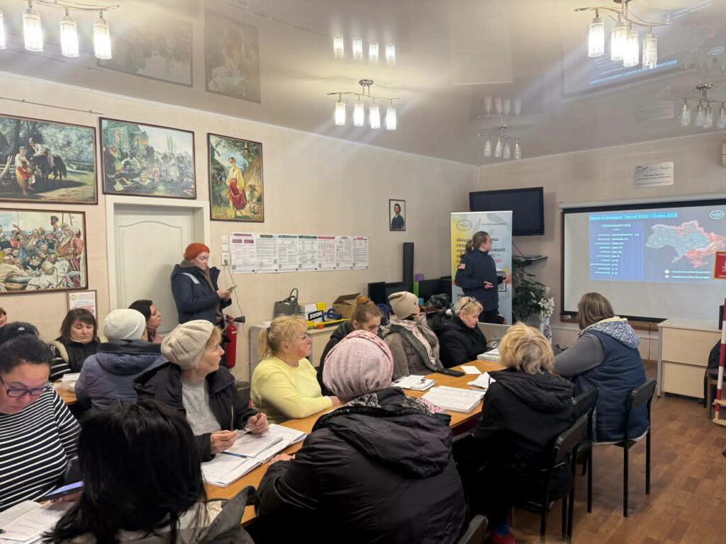 Жінка біля стіни зі світлопроводом презентує інформацію перед аудиторією, що сидить за столами.