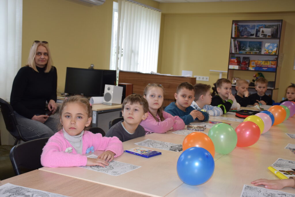 Діти сидять за столами, дивлячись вперед, біля одного з столів стоїть жінка, а на столі розкладені аркуші для розфарбовування та кольорові повітряні кульки.