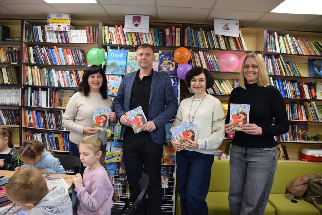 Чотири людини тримають дитячі книжки, посміхаючись на тлі книжкових полиць і дітей, що малюють.