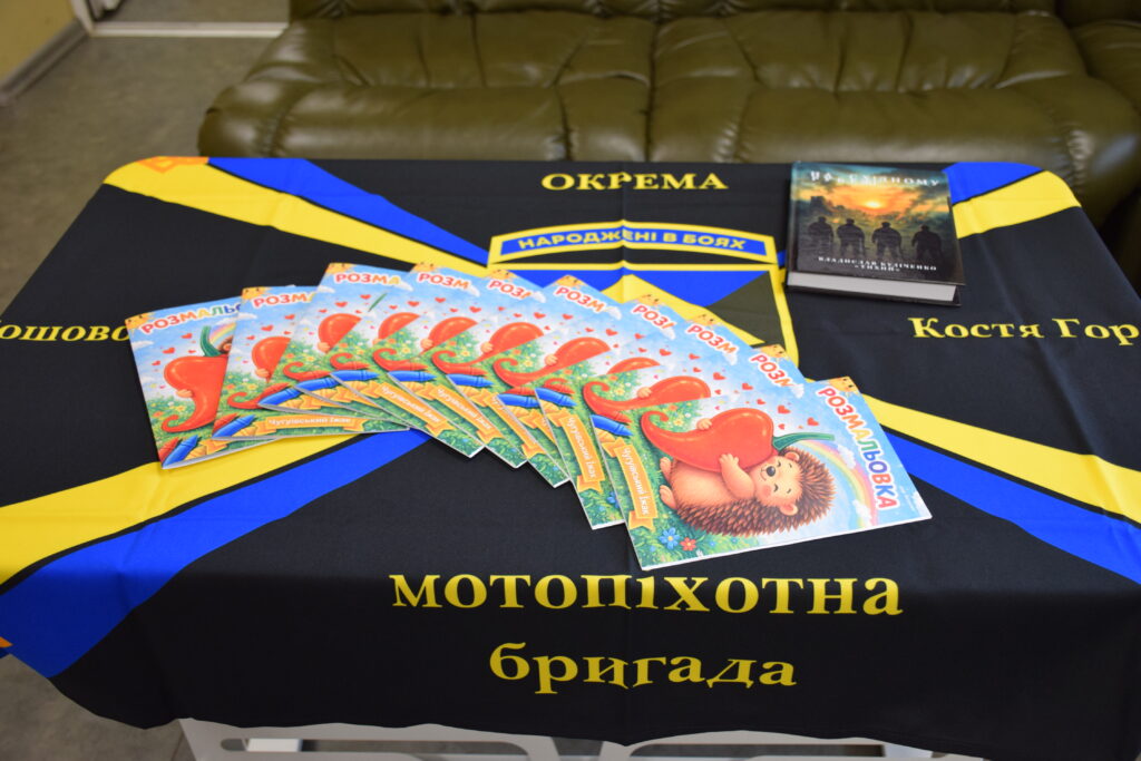 Книга з дитячою ілюстрацією та олівцями лежить на столі вкритому чорним скатертиною з написом українською мовою.