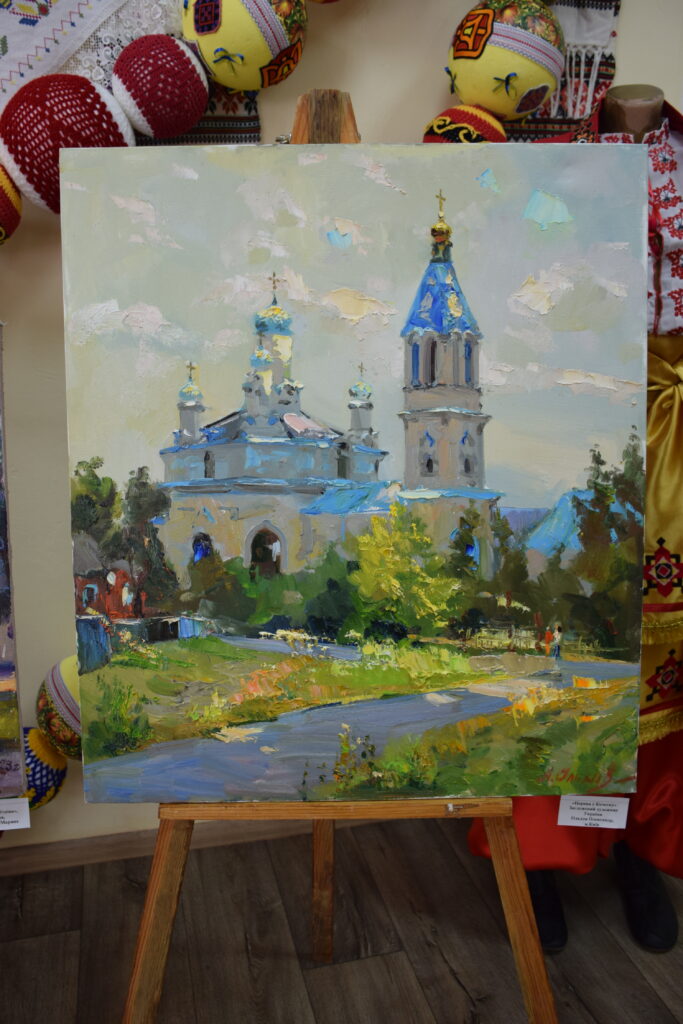 Картина на мольберті зображує церкву із синіми куполами біля річки та з написом українською мовою під малюнком.