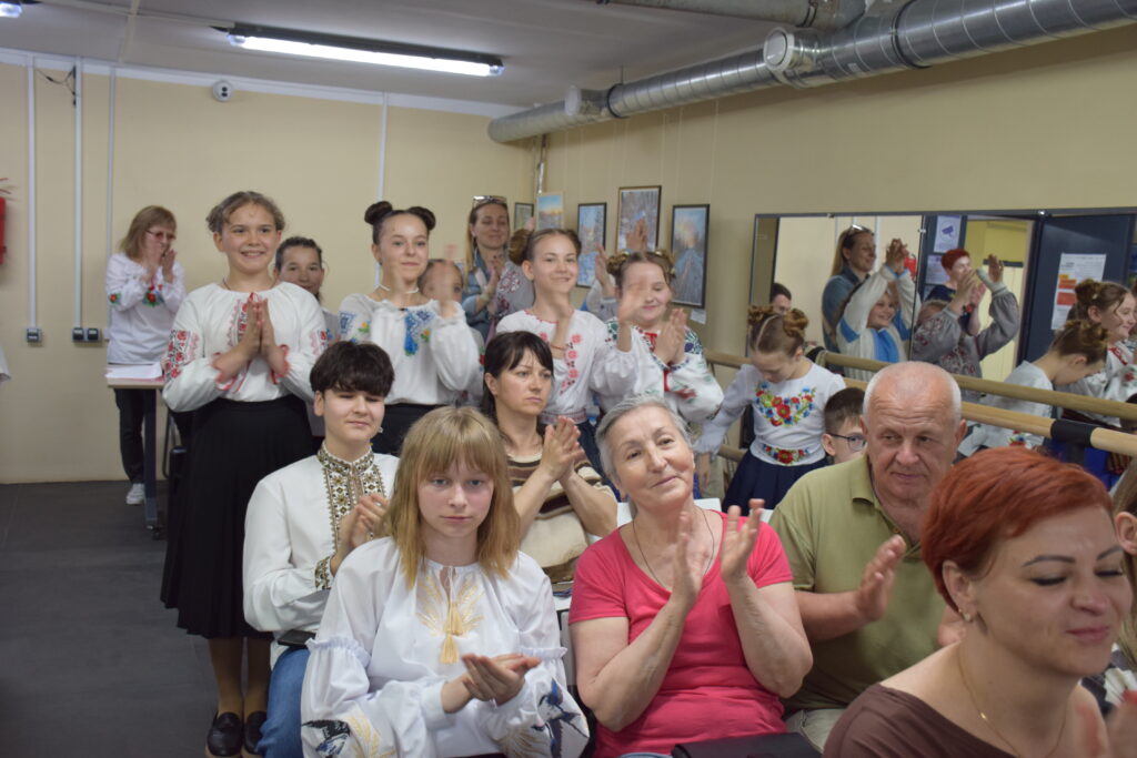 Жінка та чоловік сидять у першому ряду і аплодують разом із іншими глядачами, які стоять та сидять позаду них.