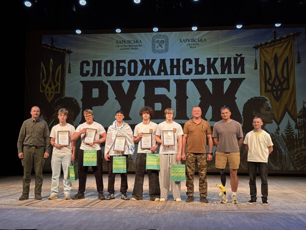 Вісім чоловік та підлітків стоять на сцені із сертифікатами та подарунковими пакетами перед великим банером із написом Слобожанський рубіж.