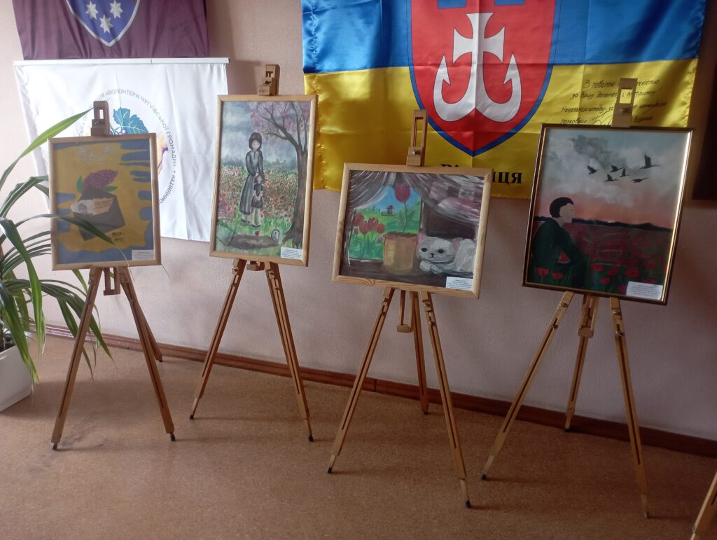 Чотири картини розміщені на підставках, встановлених перед українським прапором із гербом та іншими полотнами, що висять на стіні.