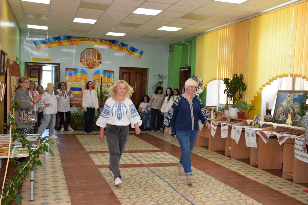 Дві жінки в українських вишиванках і джинсах ідуть коридором бібліотеки, прикрашеного вишитими рушниками та картинами, повз інших жінок, які аплодують.