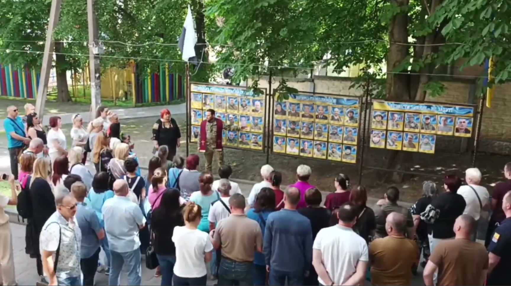 Чоловік у військовій формі виступає перед натовпом людей, що зібрались біля паркану із фотографіями.