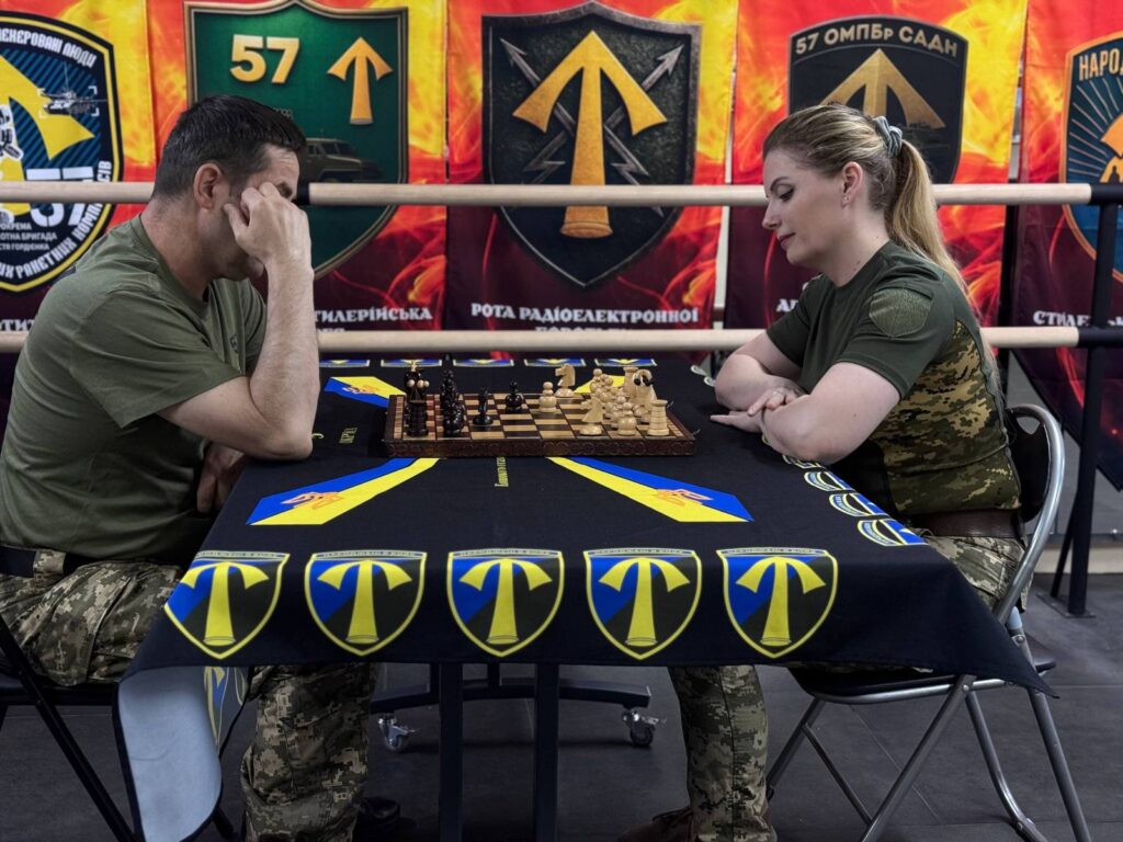 Чоловік та жінка у військовому одязі грають у шахи за столом із символічним дизайном, обличчя чоловіка виражає напруження та роздуми.