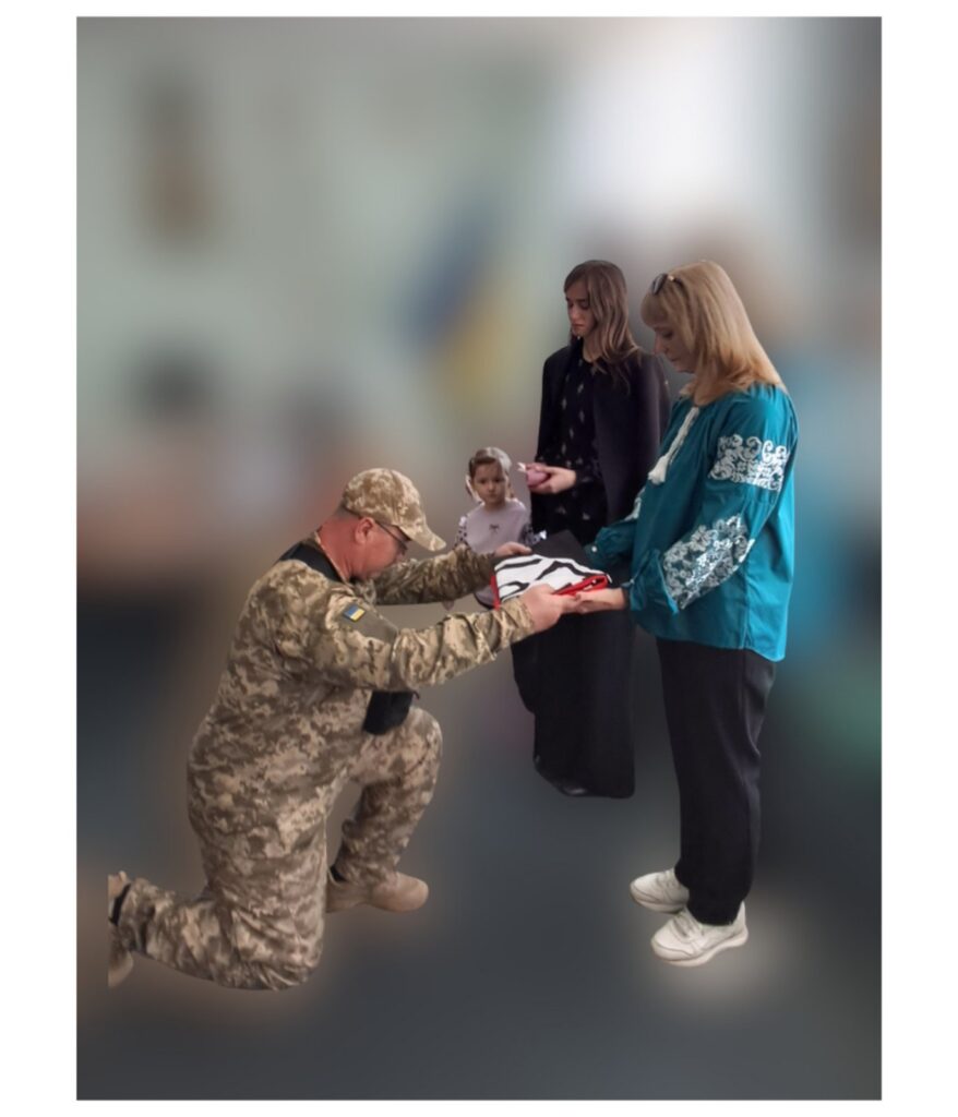 Чоловік у військовій формі на одному коліні підносить жінці пакунок, поряд стоять дві жінки та дитина.