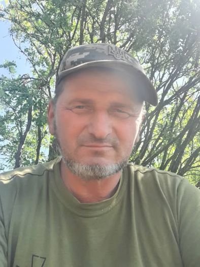 Чоловік із сівою бородою дивиться у камеру, перебуваючи на вулиці серед гілок дерев.